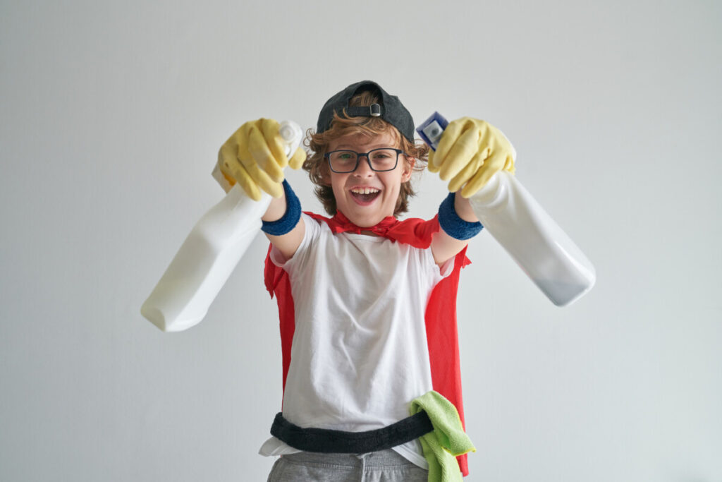 Joyful boy in housekeeper costume standing with detergent bottles in studio